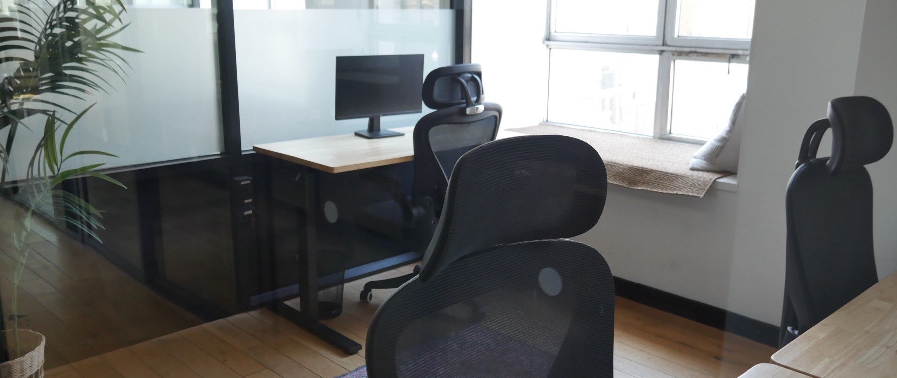 A furnished private office at Frontier Tower: a height-adjustable desk and monitor, ergonomic chairs, a cushioned window seat and wood floors, with daylight from full-height windows.
